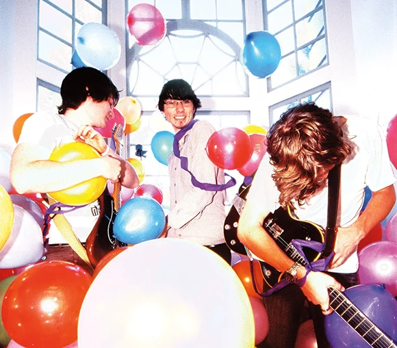 Tres músicos tocan la guitarra y la batería en una habitación bien iluminada y llena de globos.